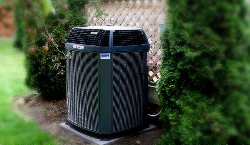 An outdoor heat pump unit set on a pad beside a garden hedge